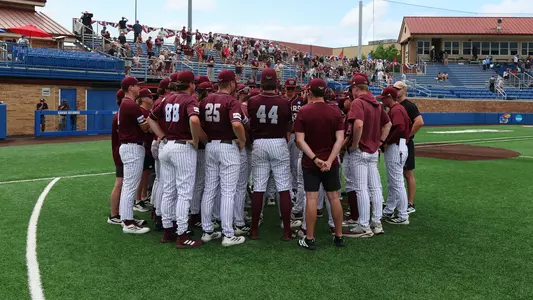 A group of baseball players in maroon jerseys and white pants stand in a huddle on a green field, with spectators in the stands behind them.
