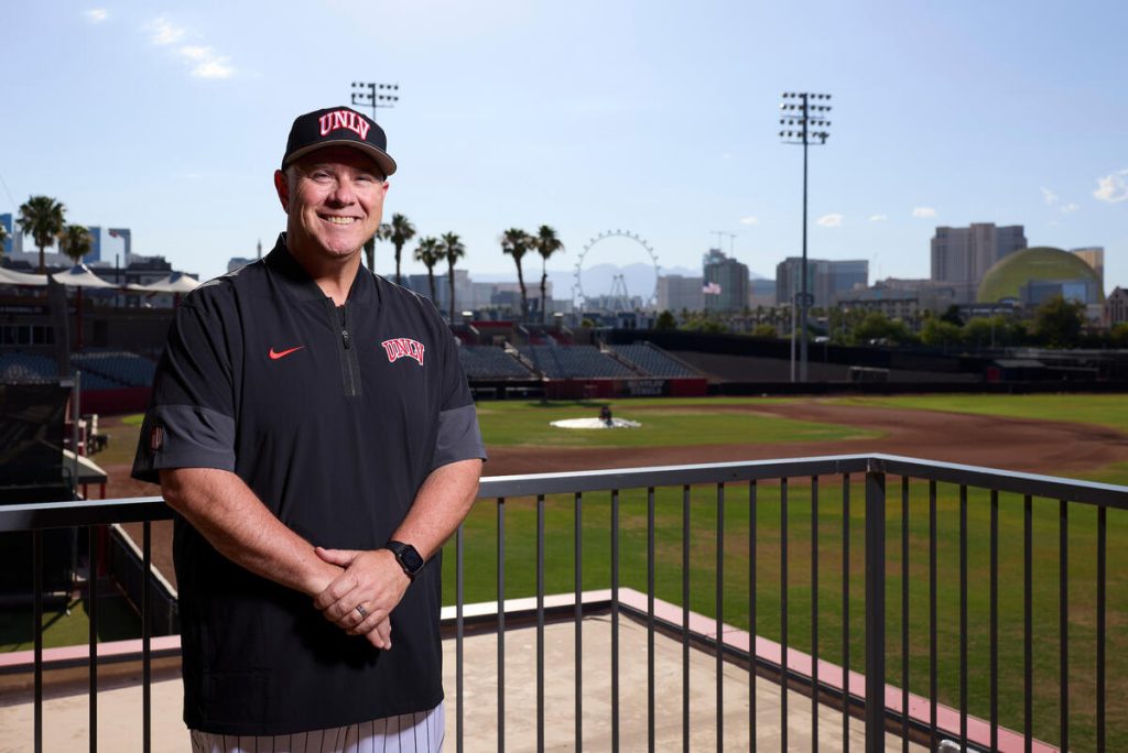 A person in a UNLV baseball uniform stands on a balcony overlooking a baseball field with a city skyline and a Ferris wheel in the background.