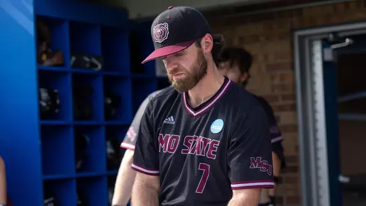 A baseball player in a Missouri State uniform stands in the dugout, looking down with a focused expression.