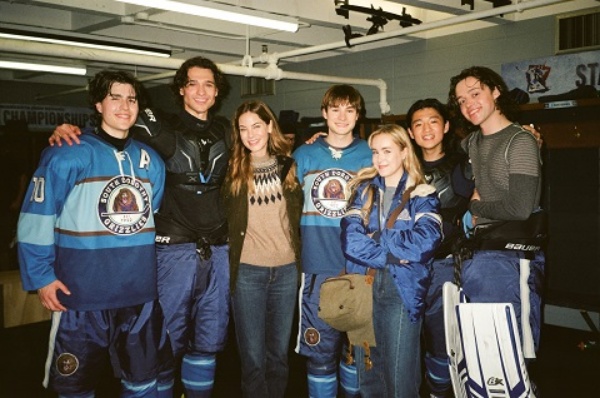 "Group of young hockey players in blue uniforms and two people in casual clothes smiling together inside a locker room, with benches and hockey equipment visible in the background."