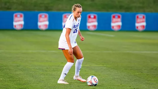 A soccer player in a white uniform with the number 28 controls the ball on a green field during a match.