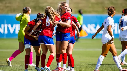 A group of soccer players in red jerseys celebrate together on a grassy field, while opponents in white jerseys walk away.