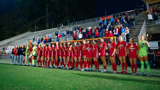 A team in red uniforms stands in a line on a soccer field at night, with spectators in the stands behind them.