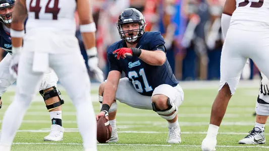 A football player in a blue jersey with the number 61 kneels on the field, holding the ball, while teammates in white uniforms stand nearby during a game.