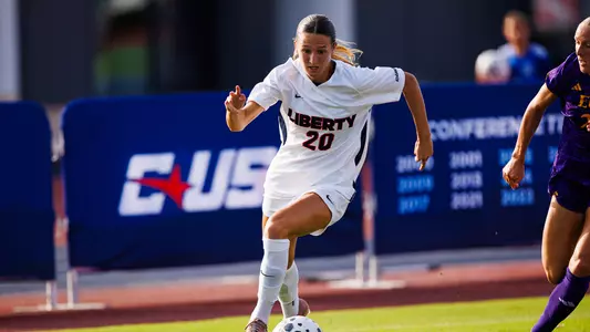 A soccer player in a white Liberty jersey with the number 20 controls the ball during a match, while an opponent in purple runs nearby.