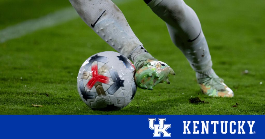 A soccer player's foot makes contact with a patterned ball on a green field.