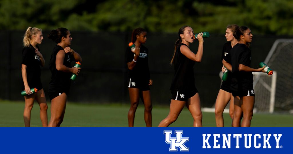 Athletes in black uniforms hydrate during a break on a grassy soccer field.