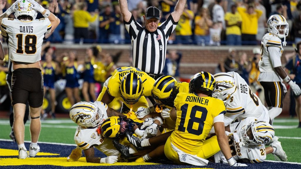 A football player in a yellow uniform is tackled by multiple opponents on the field as a referee raises his arms to signal a touchdown.