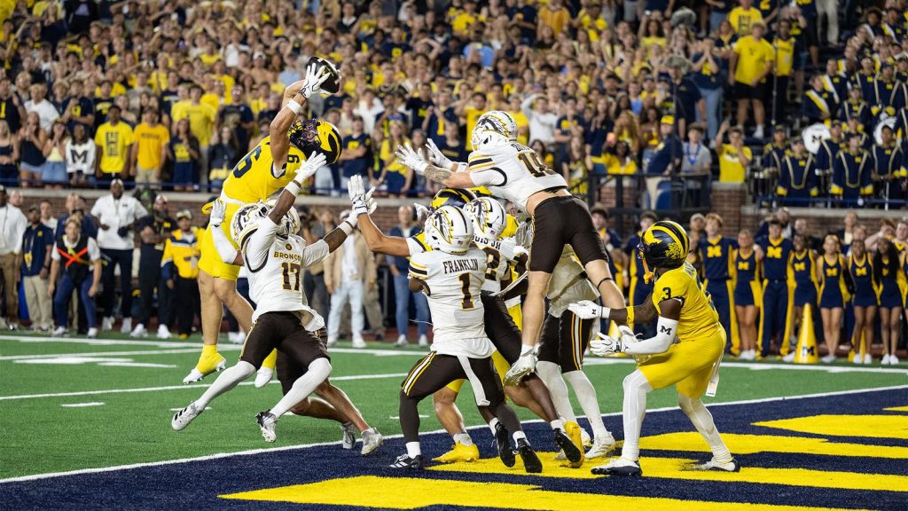 A football player in a yellow uniform leaps to catch a pass while being contested by multiple defenders in white uniforms on a field with a crowd in the background.